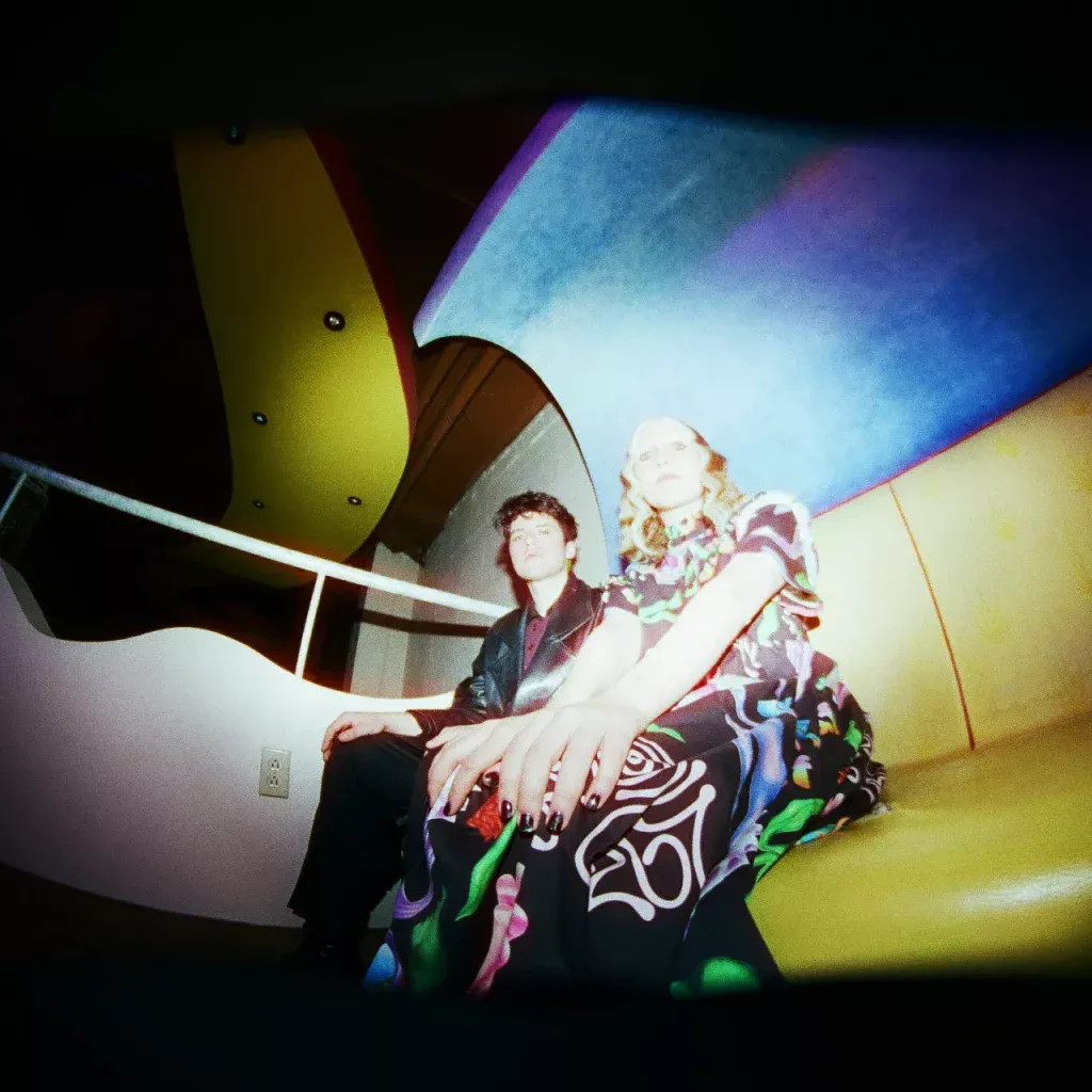 background photo of the two band members wearing dressy outfits while sitting on gold vinyl sofa with spotlight on them, centered on a receding dark background